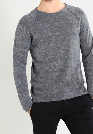 Chemise grise à manches longues en tissu doux et texturé, avec un col rond et des manches raglan, portée avec un pantalon noir.