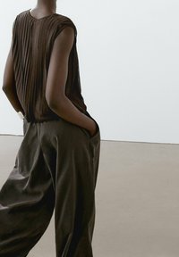 Brown pleated sleeveless top with a rounded neckline, paired with loose-fitting dark brown trousers. Smooth textures and minimal design.