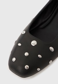 Zapato plano de cuero negro con punta cuadrada, adornado con clavos redondeados de plata de tamaños variados en la superficie superior.