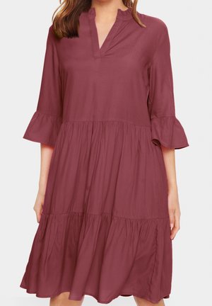 Kobieta ubrana w luźną, sięgającą kolan sukienkę w kolorze fioletowym z rękawami 3/4 rozszerzanymi ku dołowi oraz falbankowym kołnierzem w kształcie litery V.