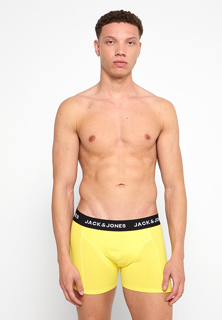 Hombre sin camiseta con cabello rizado corto, vistiendo bóxers amarillos de Jack & Jones, de pie frente a un fondo blanco.