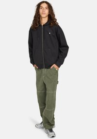 Felpa nera con zip, dotata di tasca frontale e logo, abbinata a pantaloni cargo verde sbiadito con lavorazione a patch e tasca laterale.
