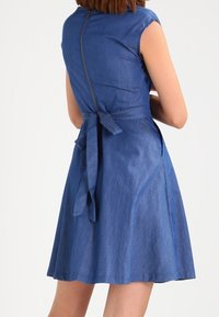 Blå denimklänning med korta ärmar, figurnära bustier, utsvängd kjol och en knytdetalj bak. Har en diskret dragkedjestängning.