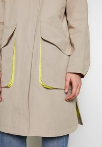 Beżowy płaszcz z dwiema dużymi kieszeniami z przodu, z jasyellow podszewką wewnątrz kieszeni. Wykonany z tkaniny o gładkiej teksturze.