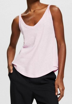 Person trägt ein hellrosa ärmelloses V-Ausschnitt-Oberteil und schwarze Hose, mit einer Hand in der Tasche vor einem schlichten Hintergrund.