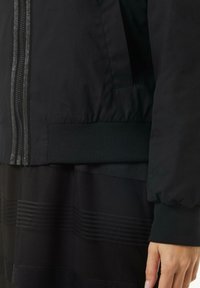 Czarna kurtka bomberka z zamkiem błyskawicznym z przodu, prążkowanymi mankietami i dolną częścią oraz gładką fakturą. W komplecie z teksturowanym czarnym ubraniem poniżej.