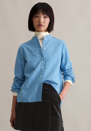 Light blue, striped button-up shirt with a band collar, long sleeves, and a relaxed fit, paired with a black corduroy skirt.