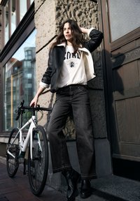 Giacca in shearling nera sopra una felpa bianca con grafica, abbinata a jeans scuri a gamba larga e stivaletti neri. Una bicicletta bianca è nelle vicinanze.