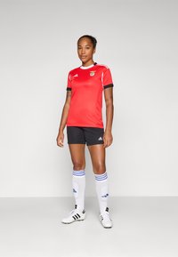 Rød Benfica fotballdrakt med korte ermer, svarte shorts og hvite sokker med blå striper, parret med hvite fotballsko.