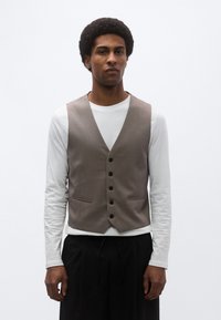 Καφέ κομψό γιλέκο με V-λαιμόκοψη, πέντε κουμπιά και πλαϊνές τσέπες, φορεμένο πάνω από λευκό πουκάμισο μακριάς μανίκας και μαύρο παντελόνι. Λευκή υφή.