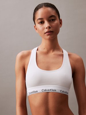 White cotton sports bra with wide straps, scoop neckline, and a branded elastic band featuring “Calvin Klein” in black. Smooth texture.