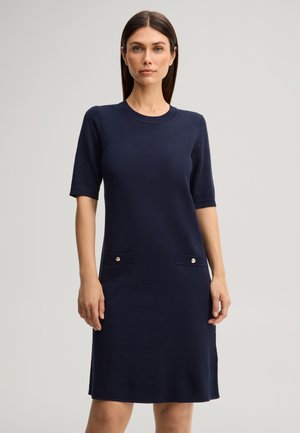 Donna che indossa un vestito blu navy lungo fino al ginocchio con maniche a gomito e due tasche frontali con dettagli a bottoni, in piedi davanti a uno sfondo semplice.