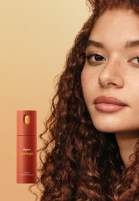 Spray Condizionante Leave-In Dyson in una bottiglia color terracotta opaco, forma cilindrica, con un pulsante giallo, accanto a un modello con capelli ricci.