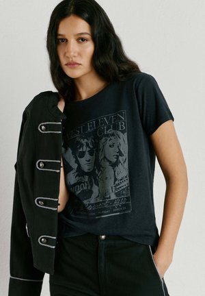 Ženska z dolgimi temnimi lasmi nosi črno grafično majico in črno jakno, prevrnjeno čez eno ramo, stoji pred enobarvnim ozadjem.