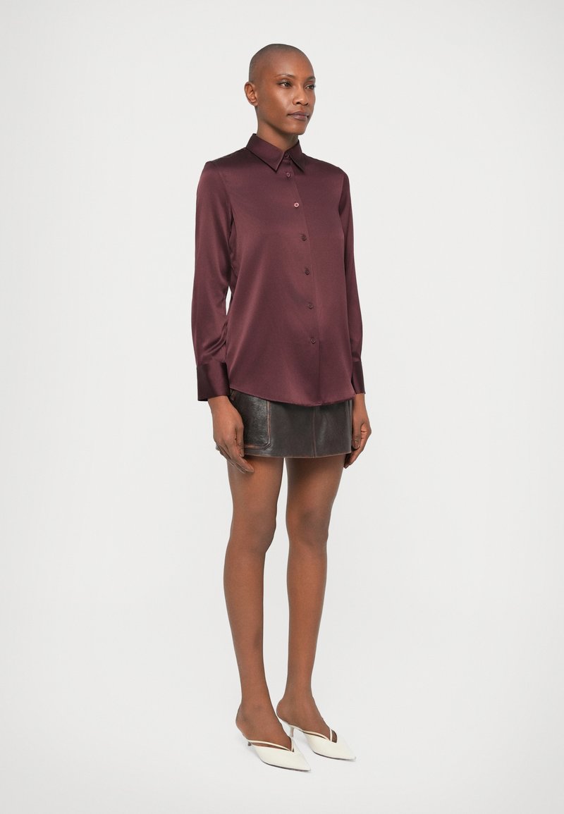 Frau steht in einem langärmeligen, burgunderfarbenen Satinhemd, einem schwarzen Leder-Minirock und weißen, spitz zulaufenden Mules vor einem schlichten Hintergrund.