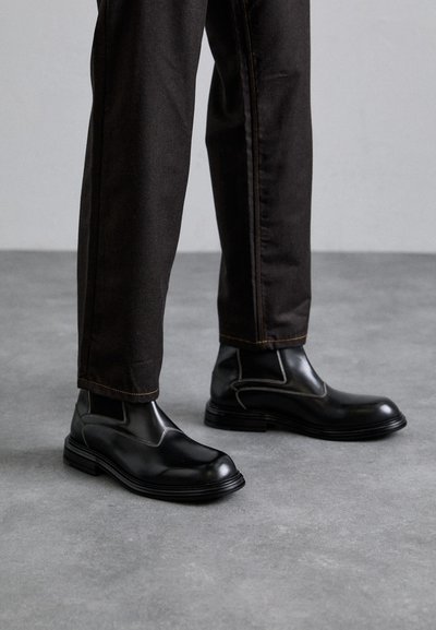 Bottines en cuir noir avec un bout arrondi, des panneaux latéraux élastiques et une épaisse semelle en caoutchouc noir, accompagnées d'un pantalon en denim foncé.