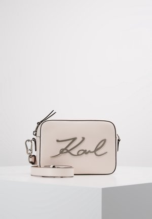 Lysebeige rektangulær crossbody-taske med sort kant, aftagelig rem og metallisk logo med kursiv skrift "Karl" på forsiden, vist på hvid overflade.