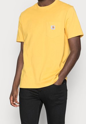 Mand iført en gul T-shirt med korte ærmer og brystlomme samt sorte bukser, hånd i lommen, mod en ensfarvet lys baggrund.