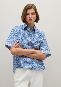 Camicia a righe blu e bianche con dettagli floreali, con bottoni sul davanti e una vestibilità rilassata. Abbinata a pantaloni bianchi semplici.