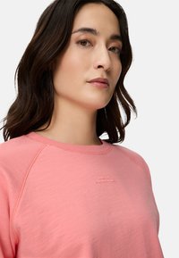 Helles rosa Sweatshirt mit Raglanärmeln, weicher Textur, dezentem Branding, rundem Ausschnitt und feinem Nahtdetail.