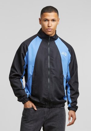 Black jacket with blue side panels, standing collar, front zip, and elastic cuffs. Features a logo on the chest and side pockets.