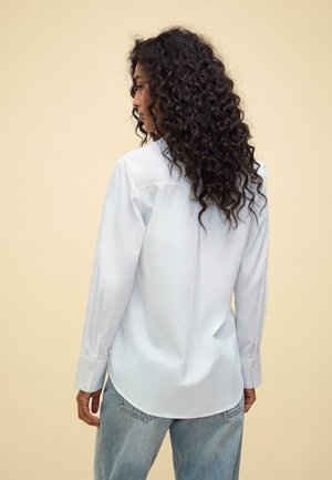 Hvid langærmet skjorte med en afslappet pasform og buet kant, parret med lyseblå denim jeans; model vist fra bagsiden.