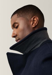 Abrigo de lana azul marino con un alto cuello gris, que presenta una forma estructurada y una tela texturada. Las líneas limpias destacan su diseño moderno.