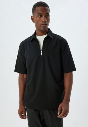 Polo noir à manches courtes avec un col zippé, fabriqué en tissu lisse, doté d'une coupe décontractée et d'un design uni.