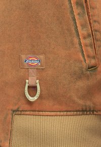 Καφέ ύφασμα κοτλέ με κυματιστή υφή. Διαθέτει ετικέτα με το λογότυπο Dickies και ένα χρυσό μεταλλικό διακοσμητικό σε σχήμα πετάλου.