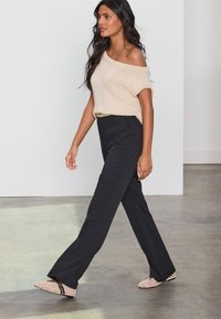 Bordinės smėlio spalvos megztas pečius atidengiantis viršus, juodos siuvinės kelnės ir šviesiai rožinės su juodais akcentais raštuotos avalynės. Modelis vaikšto neutraliame fone.