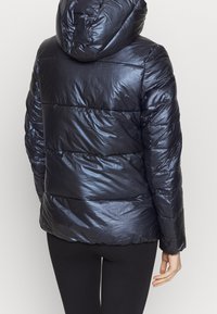 Person som bär en glänsande svart pufferjacka med huva, sedd bakifrån, tillsammans med svarta leggings mot en plain bakgrund.