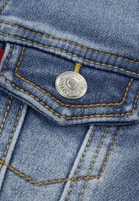 Kék farmeranyag sárga varrással, egy ezüstgombbal ellátott fedőzsebbel. Közeli részlet a textúrát és a színkontrasztot emeli ki.