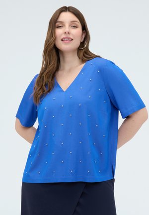 Femme portant un haut bleu à col en V avec manches courtes et petits clous argentés, associée à un pantalon foncé, debout devant un fond uni.