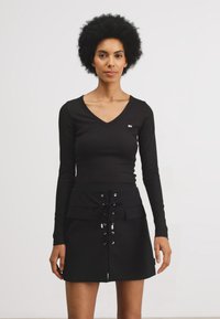 Maglietta nera a maniche lunghe con scollo a V, abbinata a una gonna nera con dettaglio di lacci frontali e due tasche frontali.