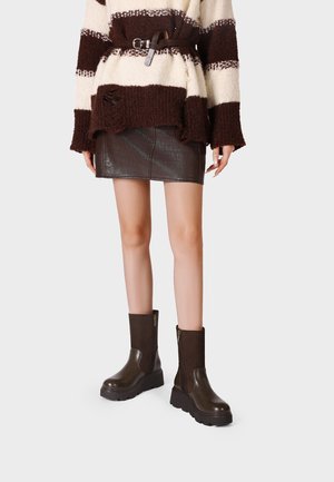 Donna che indossa una minigonna in pelle nera, un maglione a righe marrone e crema con cintura e stivaletti chunky marrone scuro con cerniere.