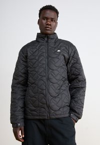 Quilts melns mētelis ar augstu apkakli, ar rāvējslēdzēju priekšā un elastīgām aprocēm. Iezīmējas ar smalku logotipu uz krūtīm. Gluda, teksturēta auduma.