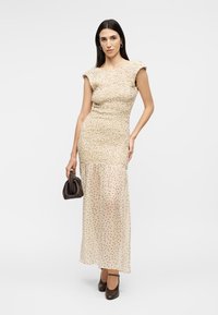 Γυναίκα που φοράει μπεζ φόρεμα με σούρες στο μπούστο και διάφανο πουά φόρεμα, κρατώντας μια μικρή καφέ τσάντα, στέκεται μπροστά σε έναν απλό φόντο.