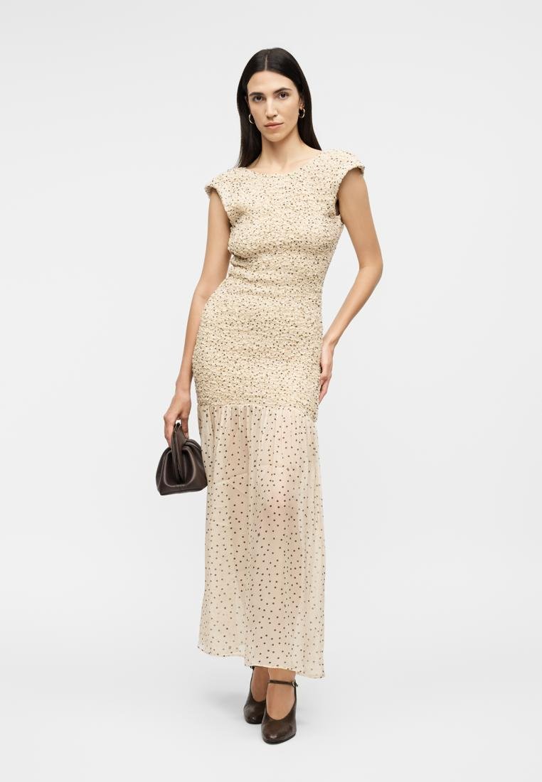 Femme portant une robe beige avec un corsage froncé et une jupe en tulle à pois, tenant un petit sac à main marron, debout devant un fond uni.