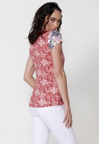 Coralfarbenes Kurzarmoberteil mit Blumenmuster, das ein weißes Blumen-Design und kontrastierende gemusterte Ärmel aufweist. Glattes Material und figurbetonte Passform.
