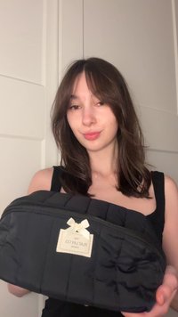 Zwarte gewatteerde cosmetische tas met een ritssluiting aan de bovenkant, een kleine witte strik als accent en een merkembleem. Zachte textuur met een gewatteerd ontwerp.