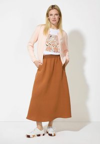 Hellrosa Strickjacke über einem weißen T-Shirt mit floralem Motiv, kombiniert mit einem braunen, fließenden Rock und weißen Sneakers mit bordauxfarbenen Akzenten.