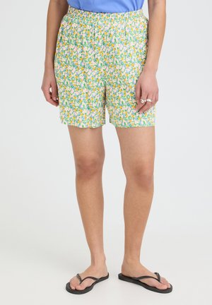 Femme portant un short à motif floral, un haut bleu, des bagues argentées et des tongs noires, debout devant un fond uni.