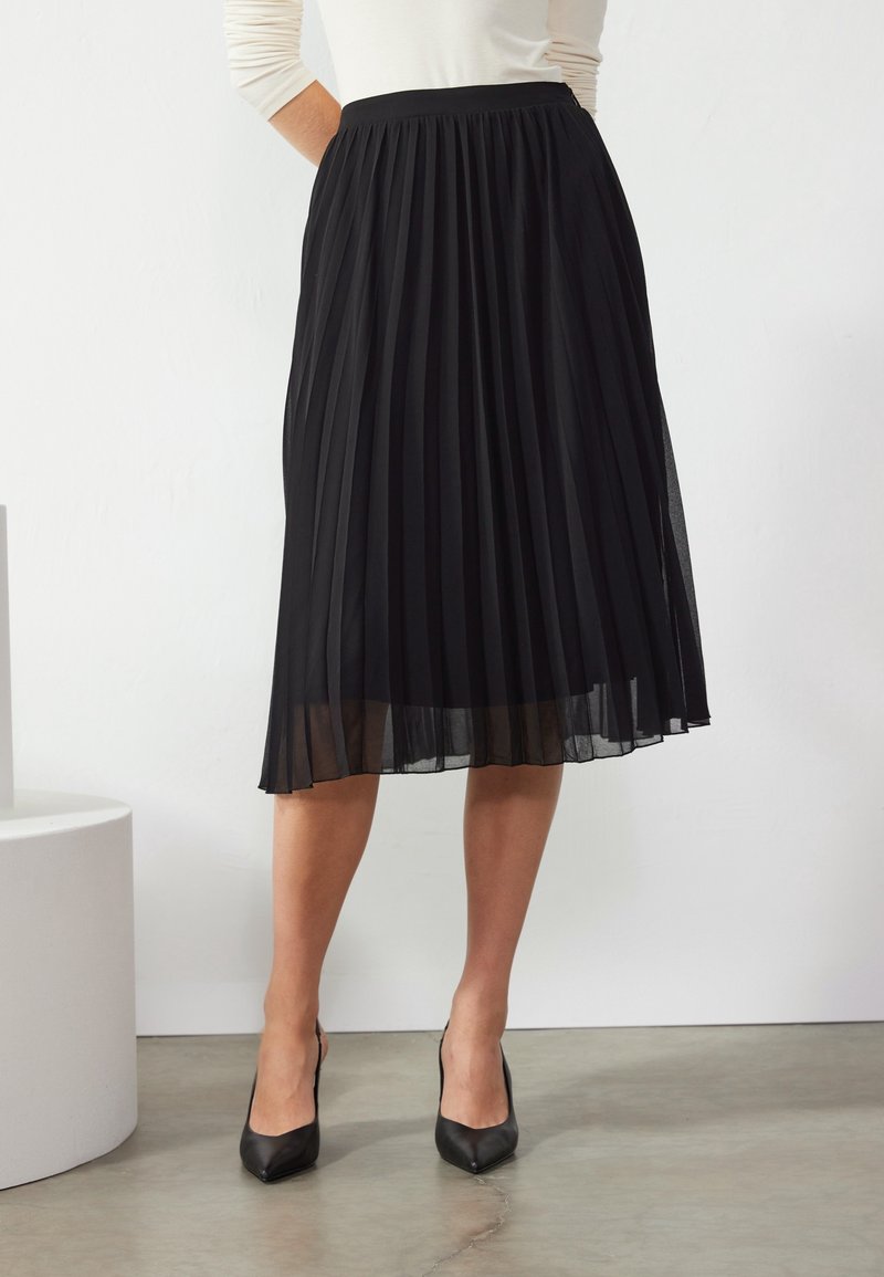 Femme portant une jupe midi plissée noire et des escarpins noirs à bout pointu, debout contre un mur blanc, les mains sur les hanches.