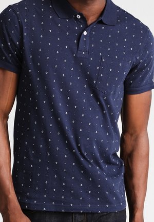 Mann trägt ein dunkelblaues Poloshirt mit kleinen weißen Anker-Mustern und einer Brusttasche, kombiniert mit einer dunklen Hose.