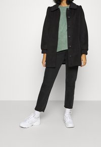 Abrigo negro de gran tamaño con cuello, botones delanteros y mangas abullonadas. Llevado sobre un suéter de punto verde y jeans negros, combinado con zapatillas blancas.