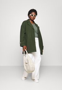 Oversized green coat, textured gray top, white wide-leg pants, white sneakers, and cream bag. Features bold sunglasses and stylish buttons.