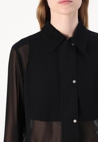 Camicia nera con bottoni, dotata di colletto e maniche lunghe trasparenti, con bottoni a pressione argento, realizzata in tessuto leggero con una texture liscia.