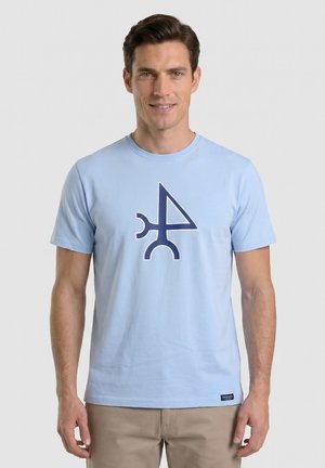 Hombre con camiseta azul clara con un símbolo geométrico abstracto azul oscuro, de pie frente a un fondo claro y liso, sonriendo.