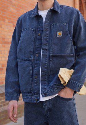 Man draagt donkerblauwe spijkerjas en spijkerbroek, witte T-shirt, houdt beige handschoenen vast, staat voor een bakstenen muur.