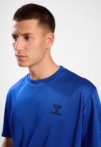 Ung man med kort hår som bär en blå hummel sporttröja, tittar åt vänster mot en enfärgad vit bakgrund.